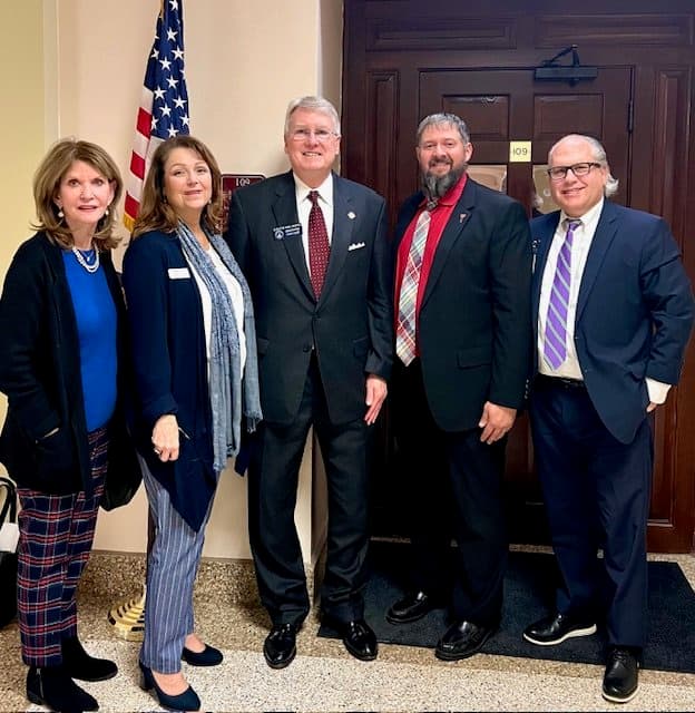 Five professionally dressed people standing together indoors near an American flag.