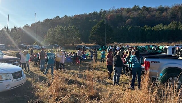 Group of people gathered outdoors in a grassy field, with cars parked nearby and trees in the background.
