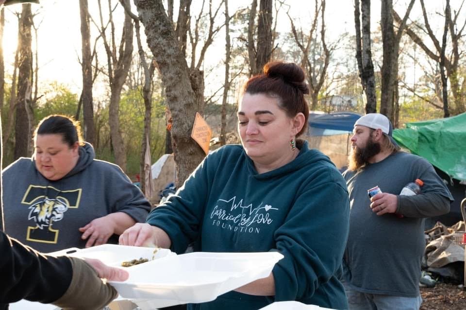 Group of three people outdoors near tents, serving food on foam plates.