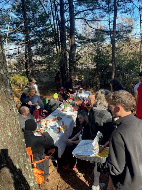 People gathered around a picnic table outdoors in a wooded area, enjoying a meal together.