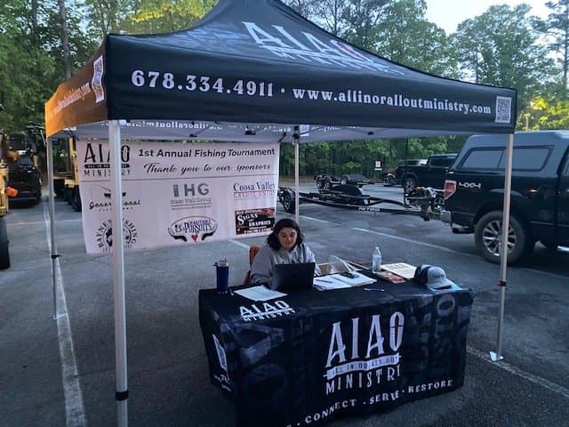 A woman at a table under a canopy tent with All In or All Out Ministry branding at a fishing tournament.