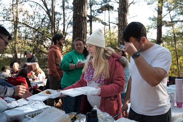 People gathered outdoors in a wooded area, serving food from a table during a community event.