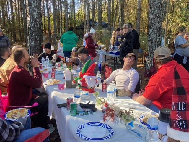 Outdoor holiday gathering in a forest with people sitting at a long table decorated with Christmas decorations.