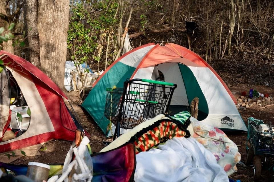 Camping scene with two tents set up in a wooded area, with camping supplies scattered around.