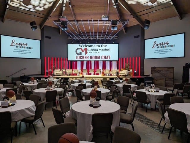 Event space set up with round tables and a stage screen reading Welcome to the Glenda Mitchell Law Firm Locker Room Chat.