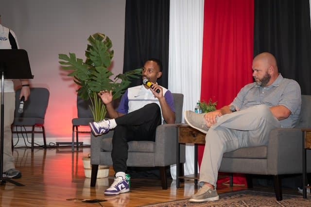 Two men seated on stage, one holding a microphone, with black, white, and red curtains in the background.