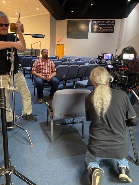 A man sitting in an empty auditorium while a crew sets up filming equipment for a podcast recording.
