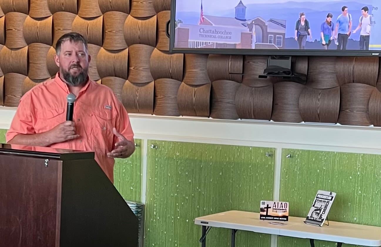 A man speaking into a microphone at a presentation, with a screen showing a campus scene.