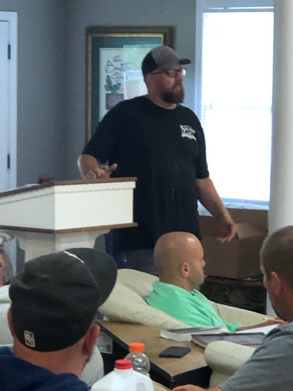 A man standing behind a table, speaking to a group seated at a meeting.