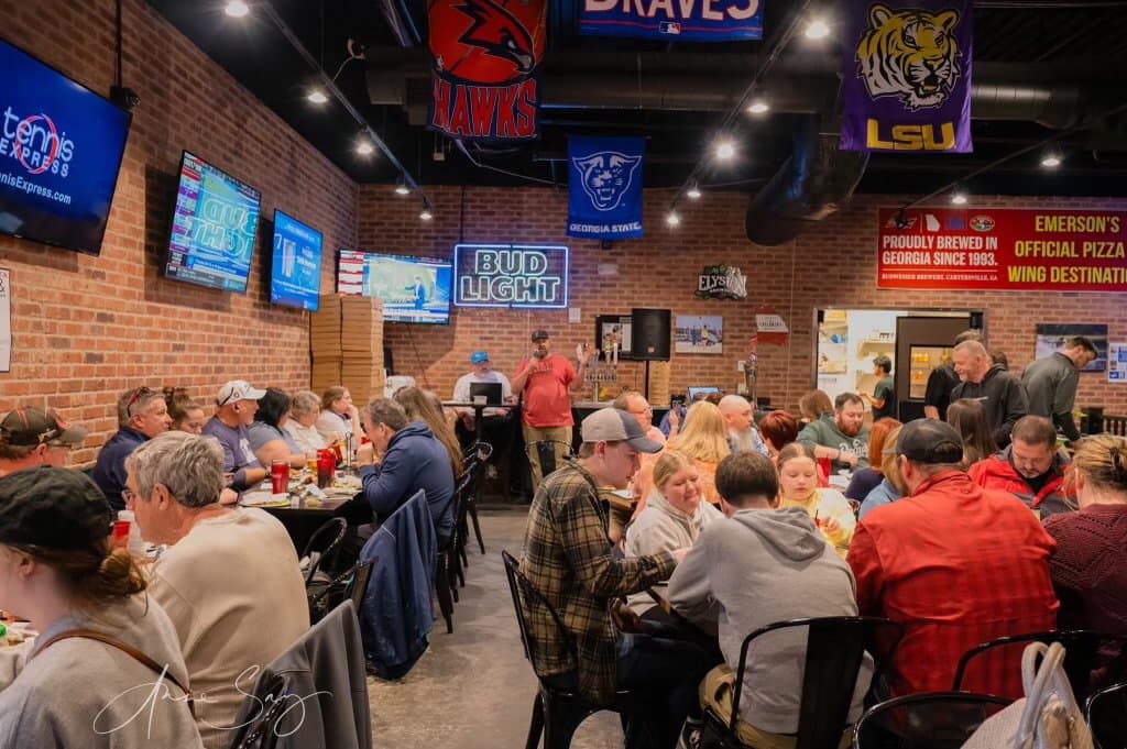A crowded restaurant with brick walls, television screens, and a DJ setup.