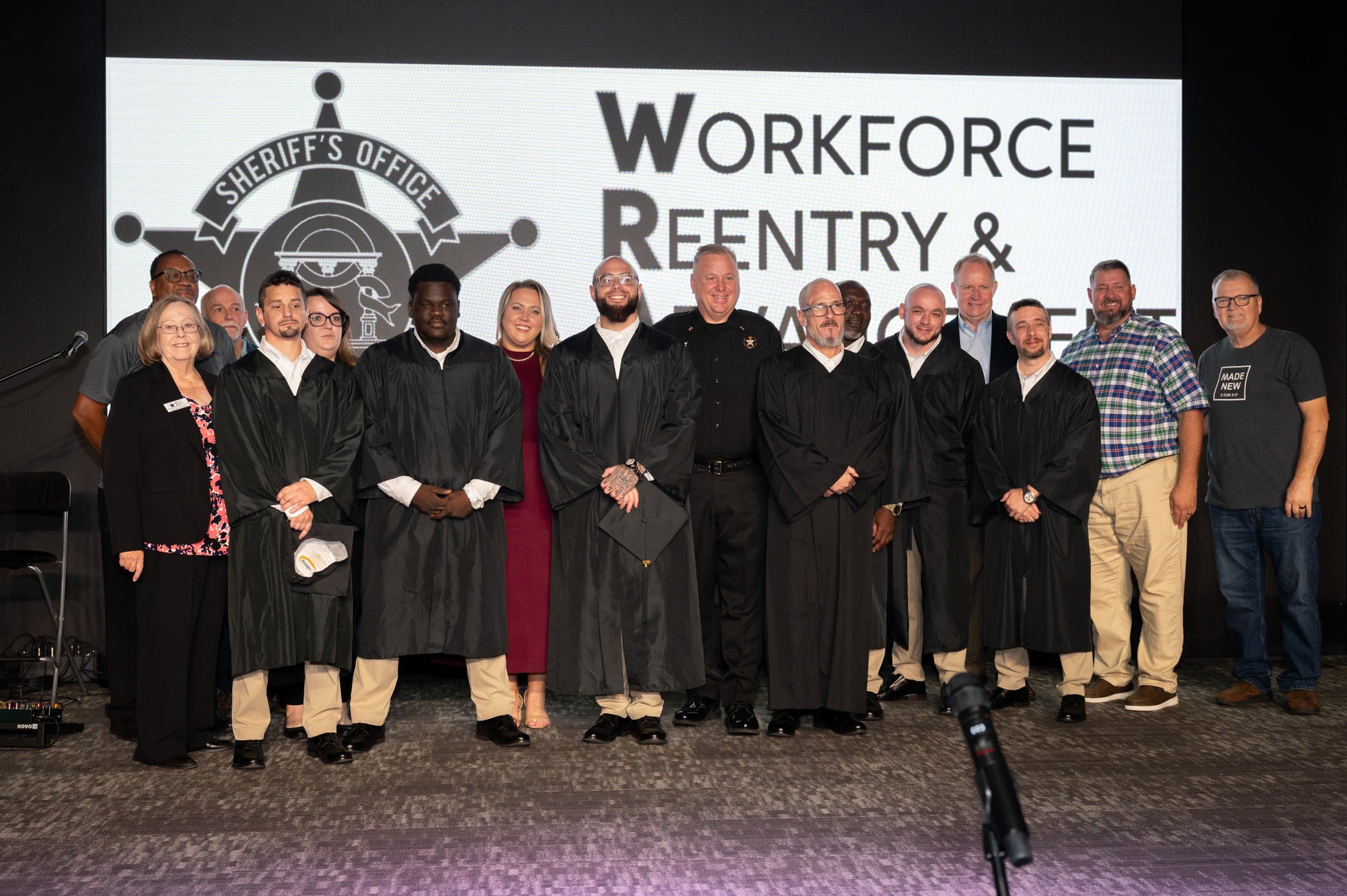 Graduates in black robes standing on stage in front of a screen displaying the Workforce Reentry & Advancement Program logo at a formal graduation event.